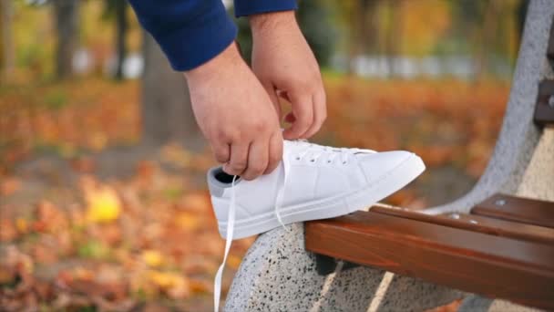 Protez bacaklı ve beyaz spor ayakkabılı bir adamın yavaş çekim görüntüsü. Ayakkabı bağcıklarını ayağıyla parkta bankta bağlıyor. Düşen sarı yapraklar - Video, Çekim