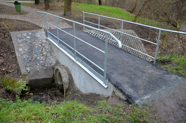 小さな小川の上の公園の新しく改装された橋。流路を通るパイプを持つコンクリート構造物です。アスファルト表面、金属板亜鉛ドリップトレイ。パイプ手すり芝生庭サイクル - 写真・画像