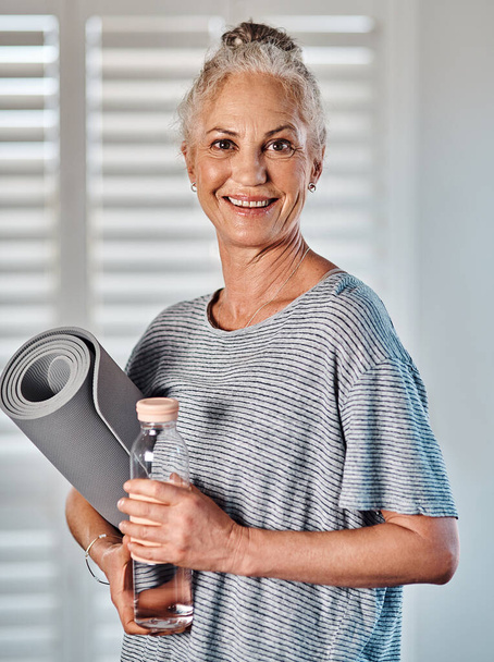 Il est temps de travailler sur ma santé et ma condition physique. Portrait d'une femme mûre joyeuse tenant une bouteille d'eau et un tapis de yoga prêt à commencer sa séance matinale de yoga à l'intérieur d'un studio - Photo, image