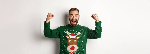 Capodanno, vacanze e feste. Uomo sollevato ed eccitato in maglione di Natale, alzando le mani e celebrando, trionfando, in piedi su sfondo bianco. - Foto, immagini