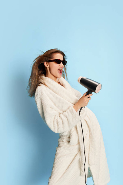 Portrait of beautiful girl in bathrobe, sunglasses posing, singing in hair dryer over blue background. Morning fun. Concept of youth, beauty, fashion, lifestyle, emotions, facial expression. Ad - Foto, Imagen