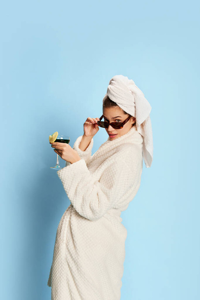 Beautiful girl in bathrobe and sunglasses, drinking cocktail over blue background. After shower. Relaxation. Party. Concept of youth, beauty, fashion, lifestyle, emotions, facial expression. Ad - Zdjęcie, obraz