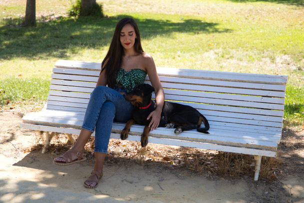 Die schöne schwarzhaarige Frau sitzt mit ihrem Dobermann-Welpen auf einer weißen Bank im Park. Die Frau genießt die Gesellschaft ihres Hundes und entspannen gemeinsam. Haustiere und Tierkonzept. - Foto, Bild