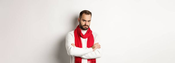 Бородатый мужчина выглядит сердитым и обиженным на тебя, скрещивает руки на груди в оборонительной позе, дуется, стоя в рождественском свитере на белом фоне. - Фото, изображение