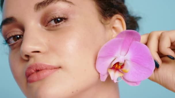 Face, flower and woman in studio for beauty, skincare and nature product, wellness and grooming on blue background. Portrait, girl and model with orchid for facial, eco friendly and floral aesthetic. - Footage, Video