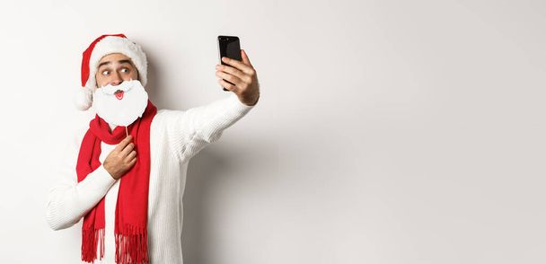 Vánoční večírek a oslavy. Mladý muž se selfie s legrační bílé vousy Santa maska a klobouk, pózování pro fotografii na mobilním telefonu, studio pozadí. - Fotografie, Obrázek