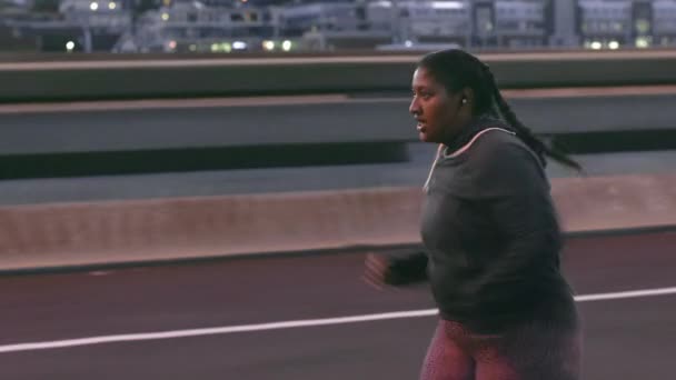 運動中のモチベーションのための音楽を聞きながら、プラスサイズの女性、ランニングや都市で夜には、ワークアウトや体重を減らすために心臓トレーニング。女性は肥満と体重減少のための都市橋で実行されます. - 映像、動画