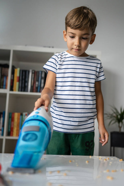 Un niño en edad preescolar limpieza desorden en la mesa con la aspiradora de mano después de jugar y hacer experimentos ayudando con la limpieza del hogar niño creciendo concepto - Foto, imagen