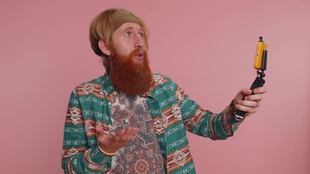 Рыжеволосый бородатый хиппи мужчина-блоггер делает селфи на смартфоне с селфи-палочкой, обмениваясь видео-звонками онлайн с подписчиками. Молодой рыжий взрослый хипстер парень на розовом фоне студии - Кадры, видео