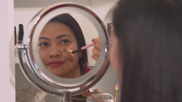 Aantrekkelijke jonge Aziatische vrouw die poeder bronzer aanbrengt om haar gezicht te verfraaien. Spiegelreflectie van een mooie dame die kwast gebruikt om gezichtsblozen aan te brengen. Vrouwelijke persoon die zorgt voor haar frisse look. - Video