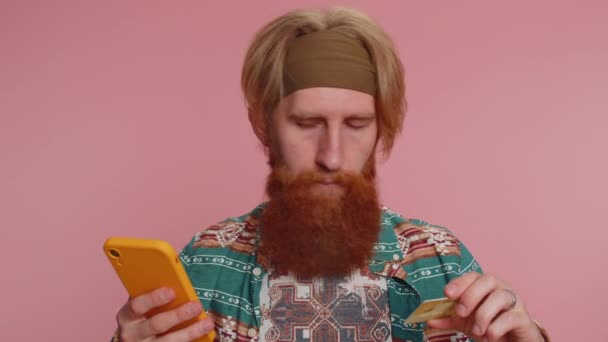 Ειλικρινής κοκκινομάλλα hippie άνθρωπος πελάτης χρησιμοποιώντας πιστωτική κάρτα τράπεζα και smartphone κατά τη μεταφορά χρημάτων αγορές online ψώνια, πληρωμή. Οικονομικά και ίντερνετ. Νεαρός απομονωμένος μόνος του σε ροζ φόντο - Πλάνα, βίντεο