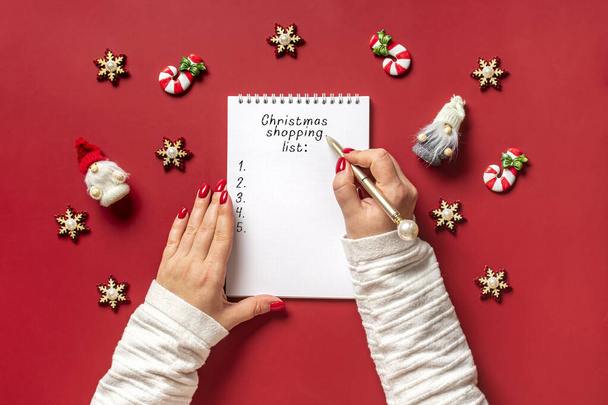 ženské ruce drží pero a psaní vánoční nákupní seznam, dárky nápady na bílém poznámkovém bloku na červeném pozadí horní pohled Flat lay Holiday card Žena dělat vánoční dárky seznam, Plánování, nákupní koncept. - Fotografie, Obrázek