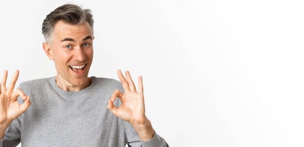 Close-up of handsome middle-aged man, showing okay signs and looking amazed, recommending something cool, standing over white background. - Foto, immagini
