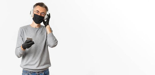 Concept van covid-19, sociale afstand en levensstijl. Beeld van man van middelbare leeftijd in medisch masker, handschoenen en bril, luistermuziek in hoofdtelefoon en smartphone. - Foto, afbeelding