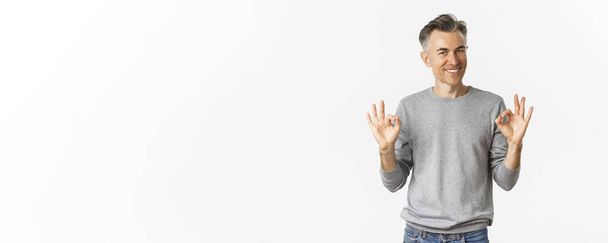 Portrait of handsome middle-aged man, smiling and looking confident while showing okay signs, guarantee something is good, standing over white background. - Фото, зображення
