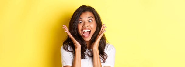 Close-up of excited african-american female model, gasping and looking amazed, raising hands up and seing something cool, standing over yellow background. - Photo, Image