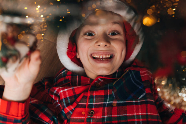 Un garçon joyeux en pyjama rouge-vert et un chapeau de Père Noël se trouve sur le sol. Noël à la maison. Vue du dessus - Photo, image