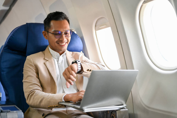 Exitoso e inteligente empresario asiático o CEO masculino se sienta en el asiento de la ventana en la clase de negocios, comprobando su reloj para la hora de llegada, en el vuelo para su viaje de negocios. - Foto, imagen
