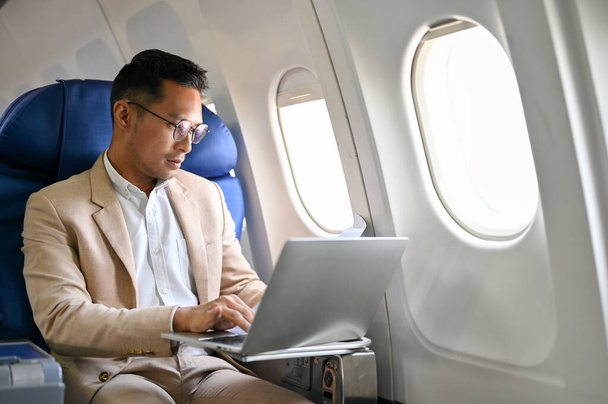 Okos és fókuszált ázsiai üzletember formális üzleti öltönyben, laptop számítógépet használ repülés közben az üzleti útjára. Üzletemberek és szállítási koncepció - Fotó, kép