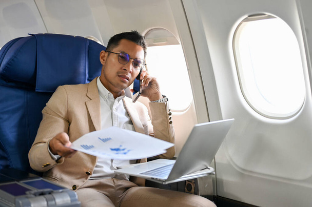 Occupé et stressé homme d'affaires asiatique a une conversation sérieuse au téléphone pendant le vol pour son voyage d'affaires. - Photo, image