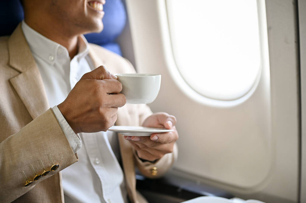 Een succesvolle en knappe Aziatische zakenman in formele kleding houdt een kopje koffie en een slokje koffie tijdens de vlucht voor een buitenlandse zakenreis. bijgesneden en close-up beeld - Foto, afbeelding