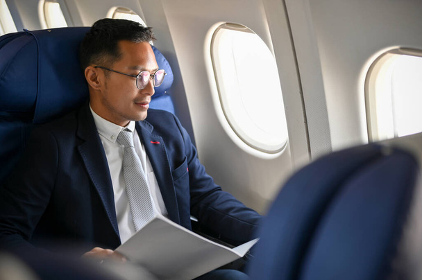 Un homme d'affaires asiatique prospère ou un PDG masculin en costume d'affaires officiel est assis à la fenêtre de la classe affaires, voyageant pour une réunion d'affaires à l'étranger. - Photo, image