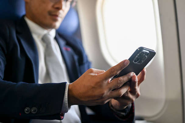 Професійний азіатський бізнесмен в офіційному костюмі сидить в бізнес-класі, використовуючи свій смартфон під час польоту. Перекручена і близькоглядна концепція - Фото, зображення