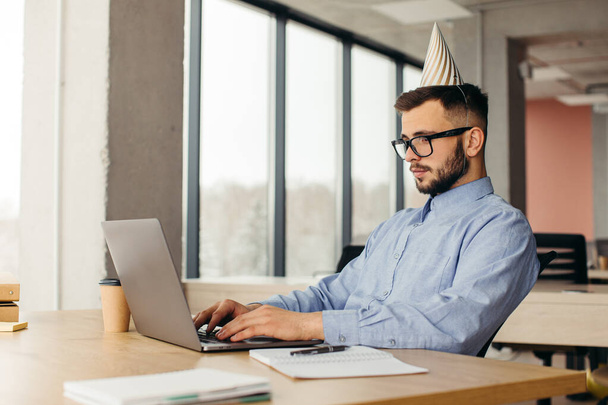 Heureux travailleur de bureau barbu positif en chapeau conique assis sur le lieu de travail avec ordinateur portable. Concept de célébration au bureau. - Photo, image