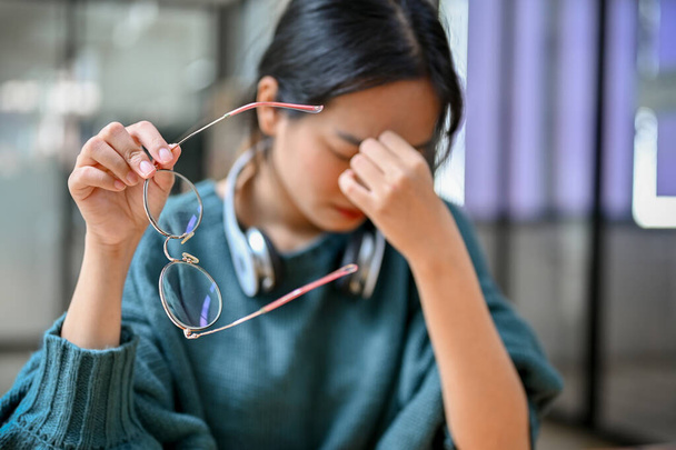 Young Asian female college student taking off her eyeglasses, suffering from eyes strain, migraine or headache while reading book. selective focus - Photo, image