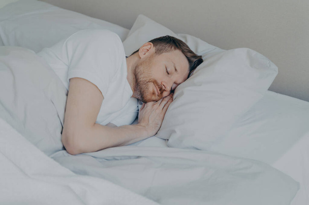 Красивый молодой человек в повседневной домашней одежде с бородой комфортно лежит и спит в уютной кровати с белыми постельными принадлежностями дома, мужчина отдыхает после тяжелого рабочего дня. Концепция сна - Фото, изображение