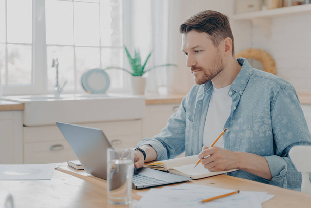 Concentrated young businessman writing notes on agenda and working remotely on laptop in internet from home while sitting at kitchen table. Freelance and work online concept - Photo, Image