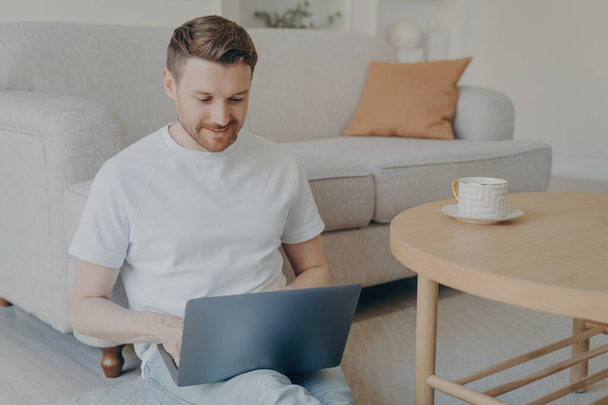 Attractive caucasian bearded freelancer man having his morning cup of coffee while sitting on carpeted floor in modern living room interior with laptop, surfing internet and working online from home - Photo, Image
