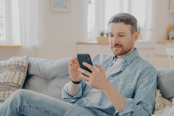 Jongeman in casual kleding surfen web op zijn telefoon en ontspannen, het controleren van berichten en meldingen tijdens het zitten op comfortabele bank met benen gekruist, wazig achtergrond - Foto, afbeelding