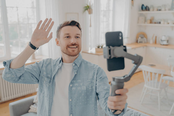 Domestic handsome guy blogger using smartphone with gimbal, waving hand in hello gesture and telling followers in social media about his life while standing in living room at home - Φωτογραφία, εικόνα