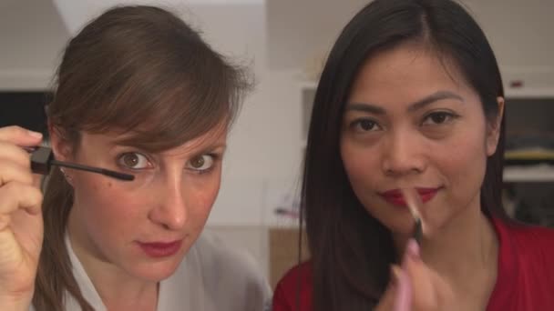 Twee dames die make-up producten gebruiken en zich klaarmaken voor een meidenavond. Aziatische en blanke vrouw die gezichtsschoonheidsproducten aanbrengt. Vrolijke vrouwen hebben plezier tijdens het doen van hun make-up. - Video