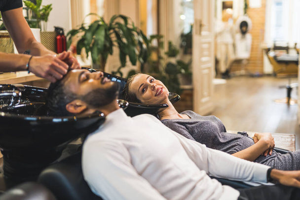 Client indien se faisant laver les cheveux par un coiffeur à côté d'une cliente blonde heureuse dans un salon de coiffure professionnel. Photo de haute qualité - Photo, image