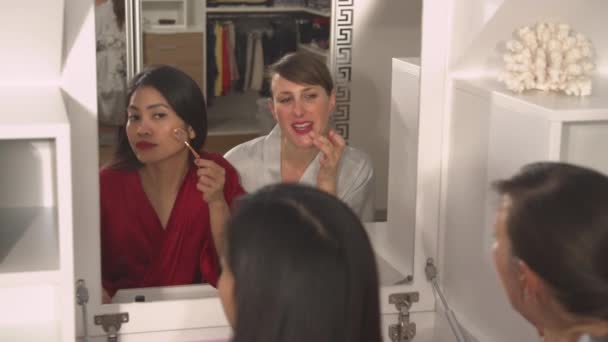 CLOSE UP: Spiegelreflectie van twee aantrekkelijke dames die hun make-up look aanbrengen. Twee mooie vrouwen die een schoonheidsbehandeling krijgen. Aziatische en blanke vrouwen die gezichtsschoonheidsproducten gebruiken voor een betere look. - Video