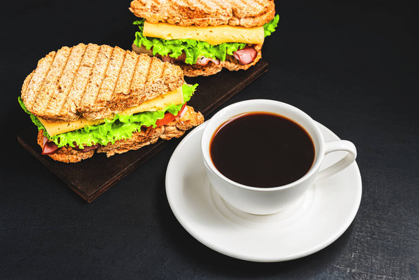 Gegrilde sandwich met kaas, sla, ham en koffiekop op donkere achtergrond. Lunchconcept met kopieerruimte. - Foto, afbeelding