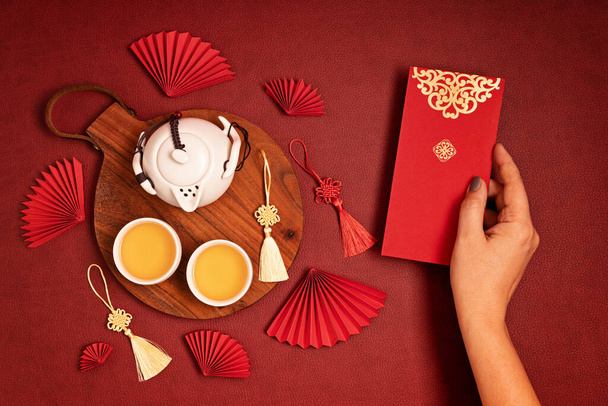 Décoration du Nouvel An chinois. Traditionnel Nouvel An lunaire plat posé avec du thé vert, ventilateurs en papier rouge, main femme tenant poche rouge. Vue du dessus - Photo, image