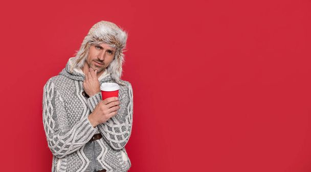 pensativo grizzled chico en suéter y sombrero sobre fondo rojo. Hoary Man bebiendo café. modelo de moda masculina mantenga taza. moda de invierno. café de la mañana en una taza para llevar. - Foto, Imagen