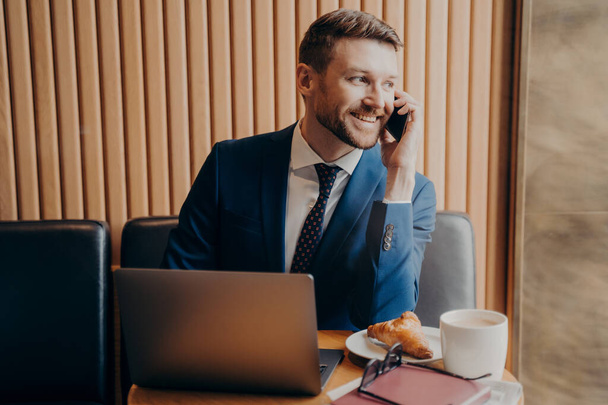 Smiling bearded handsome businessman in formal suit talking by cellphone while sitting with laptop at table in cafeteria having coffee break with cappuccino and croissant. Remote work indoor concept - Photo, Image