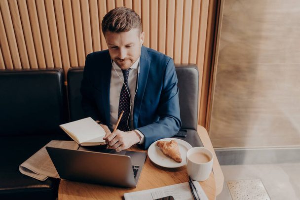 executivo de negócios masculino em terno azul em fones de ouvido estudando on-line assistindo webinar no laptop, escrevendo ideias enquanto toma café da manhã no café. Homem de negócios trabalhando remotamente em restaurante - Foto, Imagem