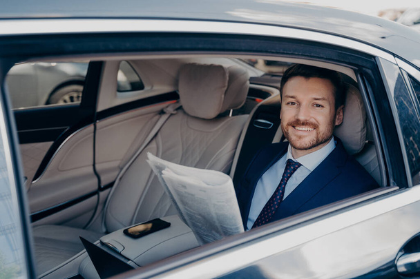 Jeune propriétaire d'entreprise indépendant souriant en costume bleu, assis à l'arrière de la limousine, vérifier le journal, heureux de lire les bonnes nouvelles écrites sur lui-même, faire un tour à l'arrière de la voiture - Photo, image