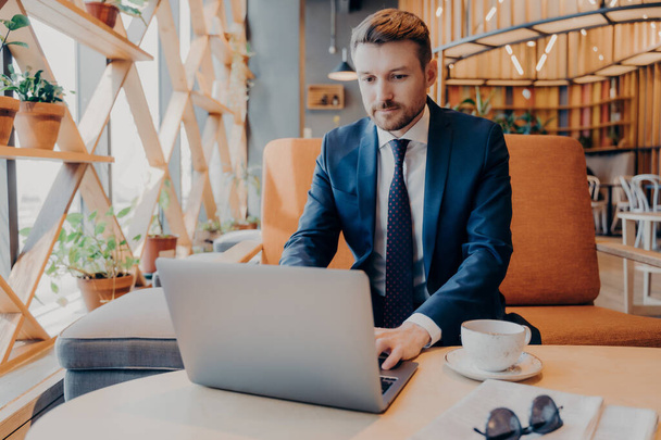 Молодой уверенный бизнесмен в формальном костюме работает онлайн на ноутбуке в лобби корпоративного центра во время кофе-брейка, готовясь к презентации для деловой встречи с новыми партнерами - Фото, изображение