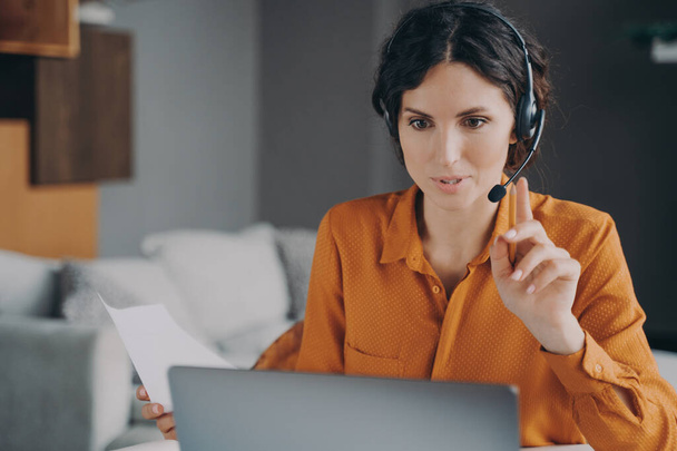 Junge Spanierin mit Kopfhörer, die während des Online-Unterrichts auf den Laptop schaut, während sie zu Hause aus der Ferne arbeitet, Callcenter-Betreiberin berät Klientin, während sie an ihrem Arbeitsplatz sitzt - Foto, Bild