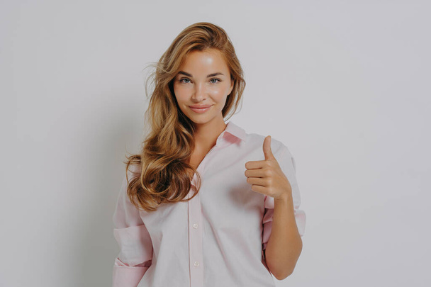Enthusiastic young Caucasian woman keeps thumb up recommends something very good says yes gestures indoor dressed in pink shirt isolated over grey background shows positive feedback. I like it - Photo, Image