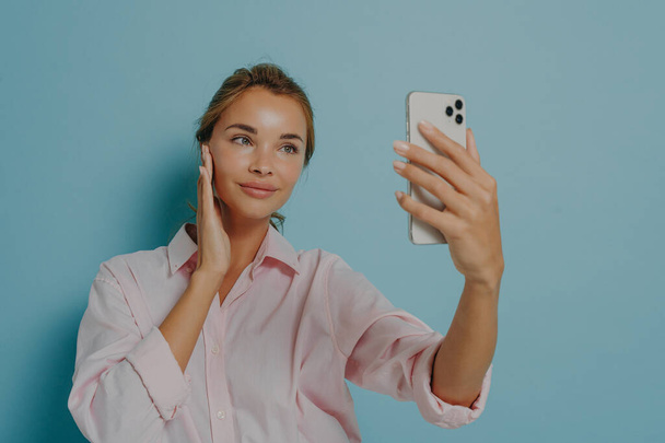 スマートフォンのカメラを見ていると、優しい表情の美しい女性は、ビデオアプリ上の青い背景トークの上に隔離された正式なシャツを身に着けています。通信とオンライン技術 - 写真・画像