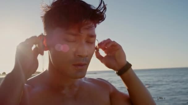 Beach, rest and man take off earphones after running, exercise or workout. Music, sports and face of exhausted male runner from India on break at sunrise after jog, training or exercising by seashore. - Кадри, відео