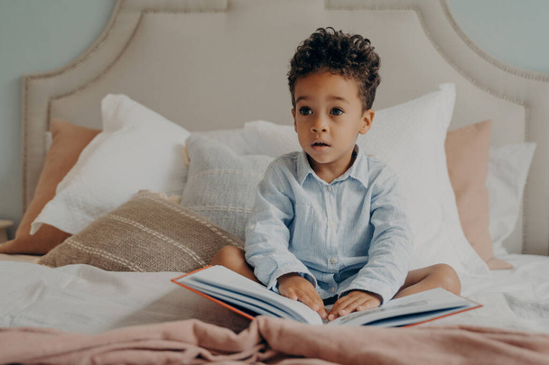Precioso niño afro americano de pelo rizado con ropa casual sentado en la cama lleno de almohadas con libro de cuentos en su regazo en casa, mirando a un lado con expresión de cara curiosa. Niño jugando en interiores - Foto, Imagen
