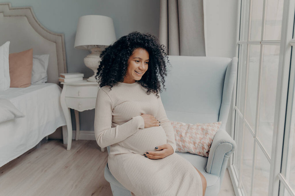 Charming young mixed race pregnant woman, future mother looking out of big window and dreaming about maternity while sitting sideways on armchair in bedroom, holding belly with both hands and smiling - Photo, Image